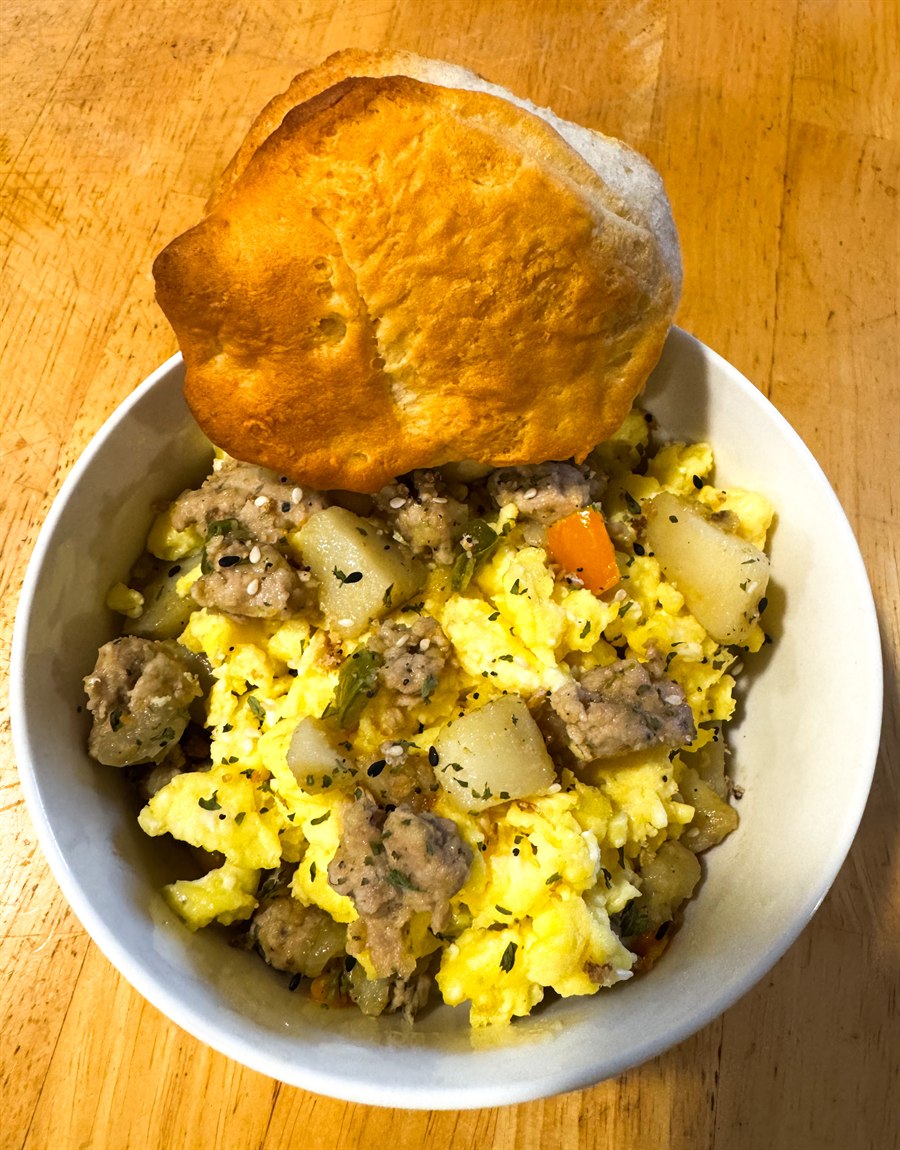 A bowl of turkey sausage breakfast hash with potatoes, peppers and scrambled egg, with a biscuit on top